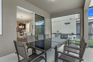 A modern balcony with a glass table and chairs, a sliding glass door leading to a dining area, and a view of the neighborhood.
