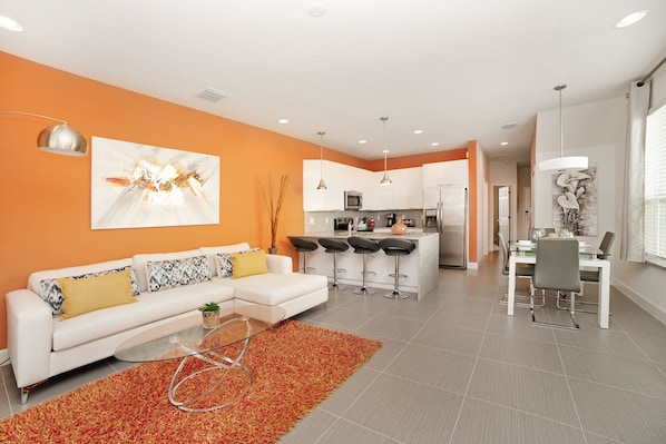 A modern living room with a white sofa, a glass coffee table, and a vibrant orange rug. A kitchen with white cabinets and a dining area with a white table and chairs are visible in the background.