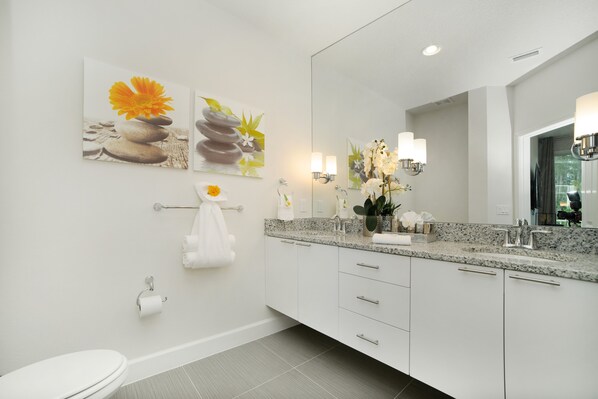 A modern bathroom with a granite countertop, white cabinets, a large mirror, and artwork on the wall.