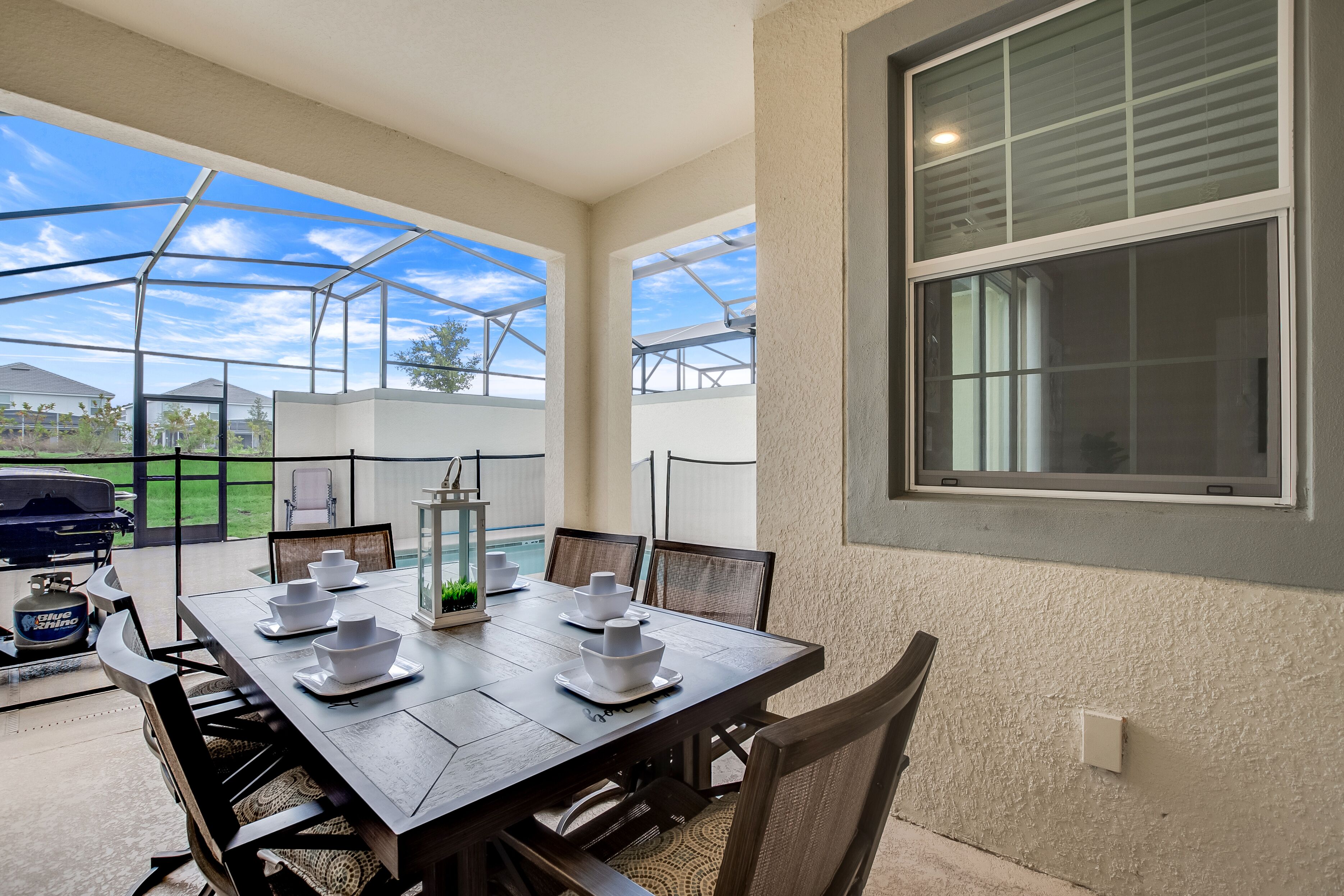 A covered patio with a dining table set for four, surrounded by chairs, and a grill in the background.