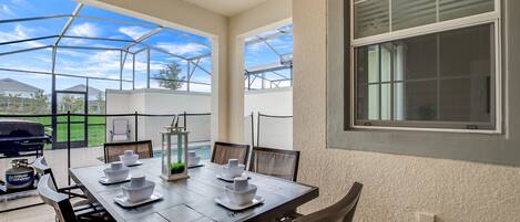 A covered patio with a dining table set for four, surrounded by chairs, and a grill in the background.
