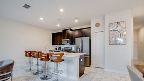 A modern kitchen with dark cabinets, a white island, and stainless steel appliances. There are four stools around the island and a chair in the foreground. - 4 Bed 3 Ba Champions Gate Pool Home (Davenport)