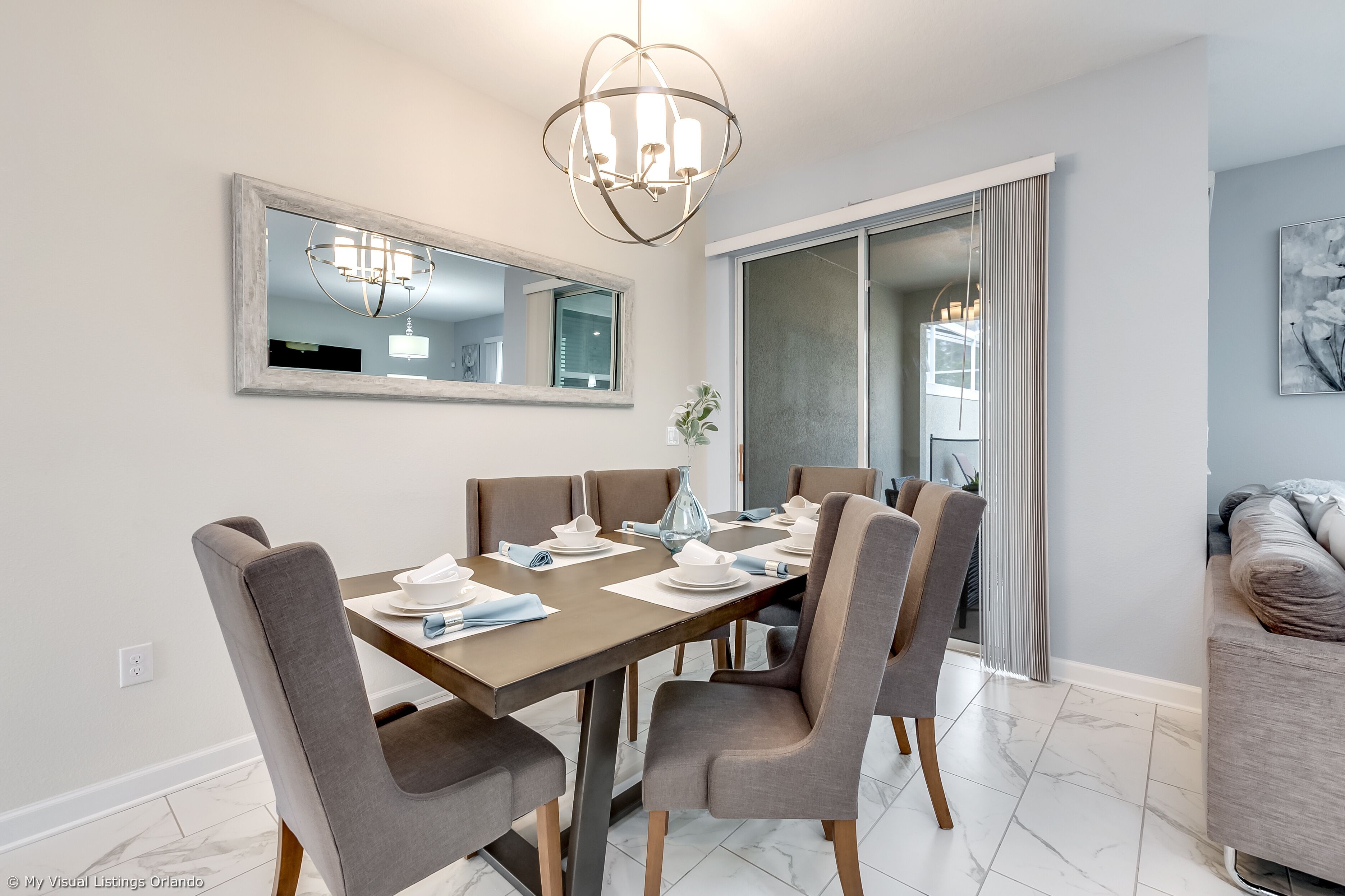 A dining area with a rectangular table, six chairs, a chandelier, a large mirror, and a sliding glass door leading to a balcony.