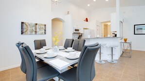 A modern dining area with a white table set for six, grey chairs, and a vase of yellow flowers. A kitchen is visible in the background with white cabinets and a microwave. - 2521rose (Davenport)
