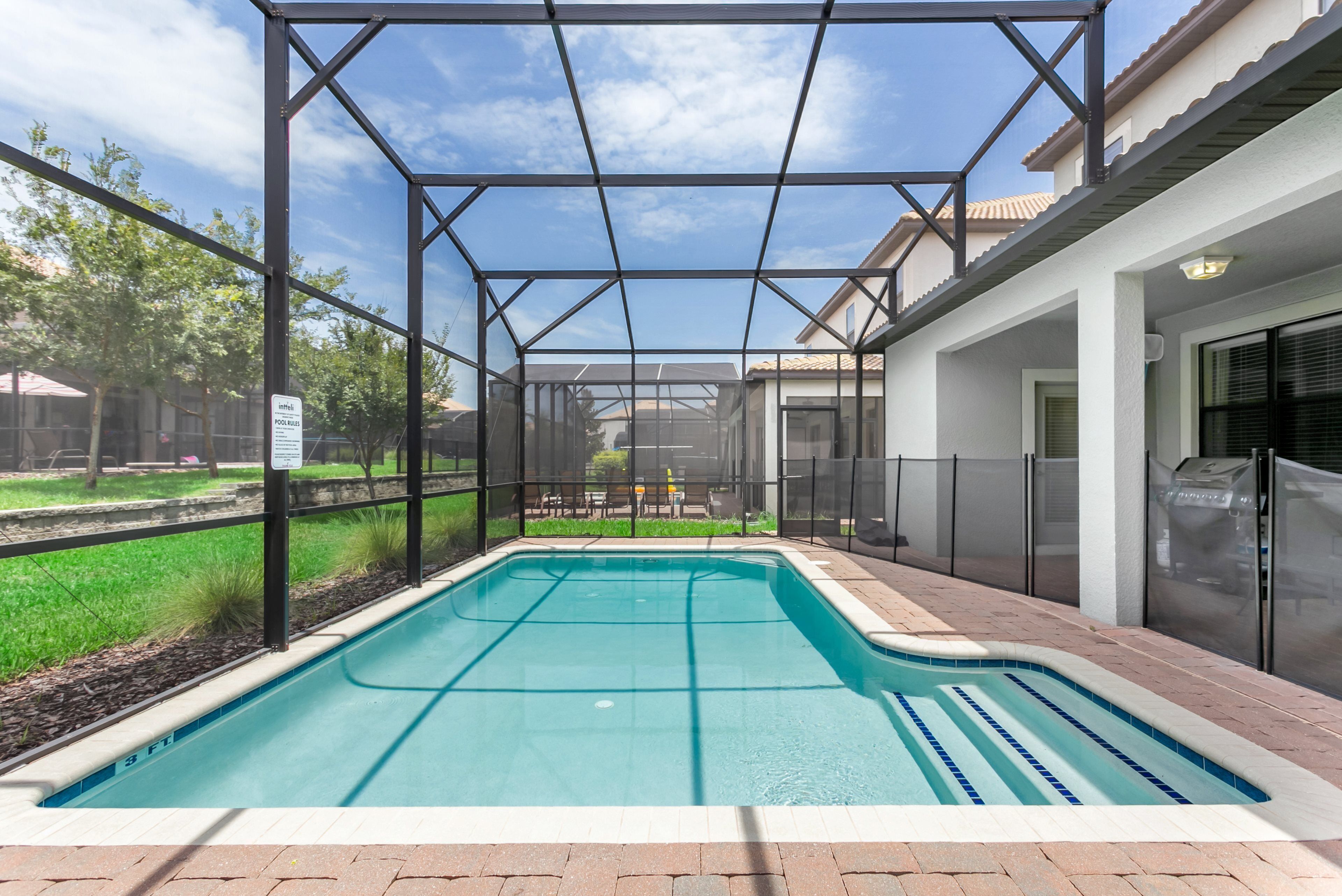 A swimming pool with a metal frame and glass panels, surrounded by a brick patio and a grassy area.