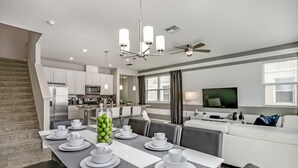 Une cuisine moderne équipée d’appareils électroménagers en acier inoxydable, un coin repas avec une table grise pour six personnes et un salon avec un canapé blanc et un téléviseur à écran plat.