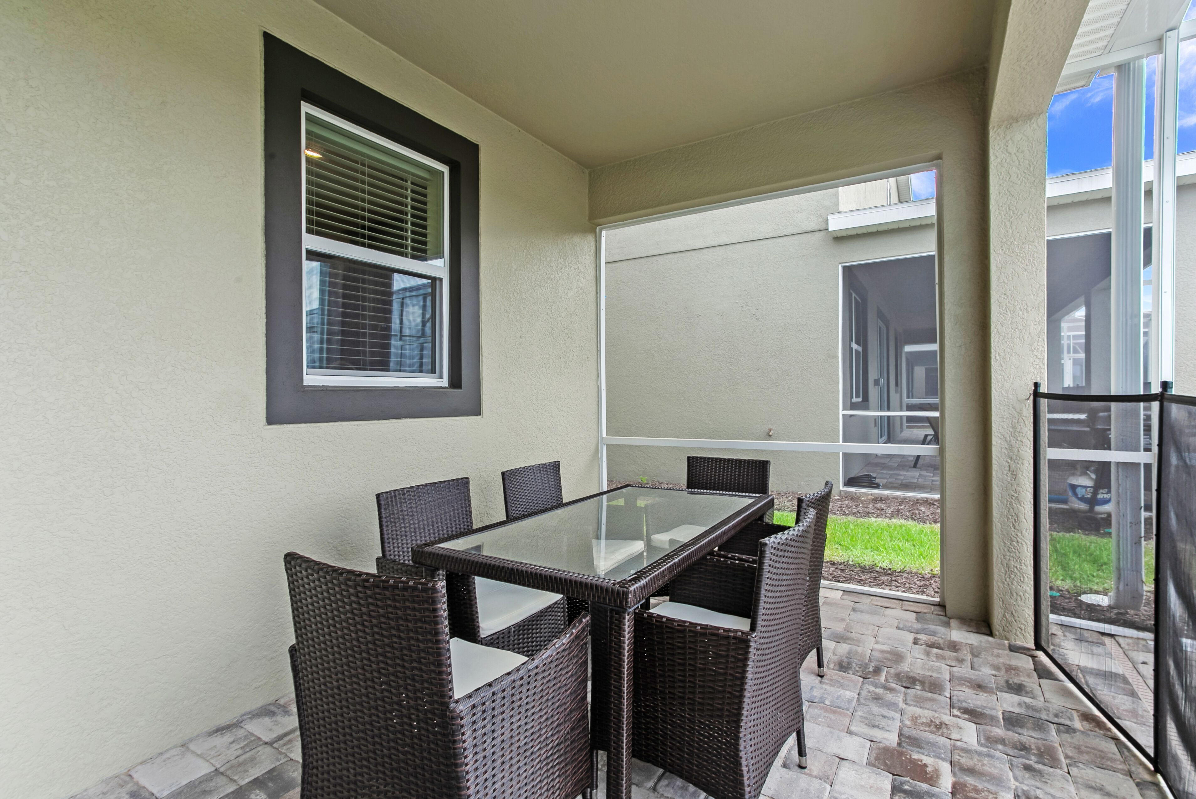 A patio area with a glass-top table and wicker chairs, a window with blinds, and a view of a house through a glass door.
