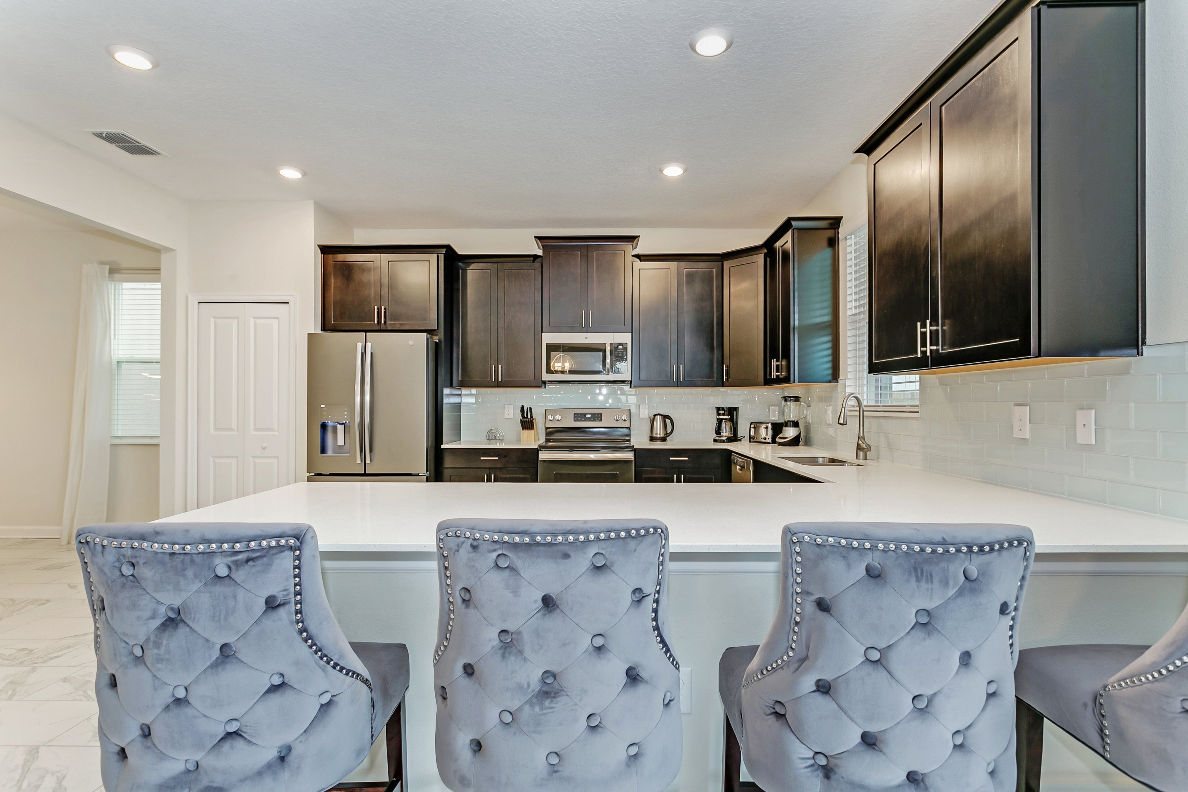 A modern kitchen with dark cabinets, a white countertop, and three tufted chairs.