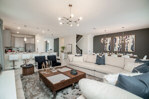 Un salon moderne avec un canapé, une table basse et des fauteuils. On aperçoit une cuisine à l’arrière-plan, avec un réfrigérateur et des placards.