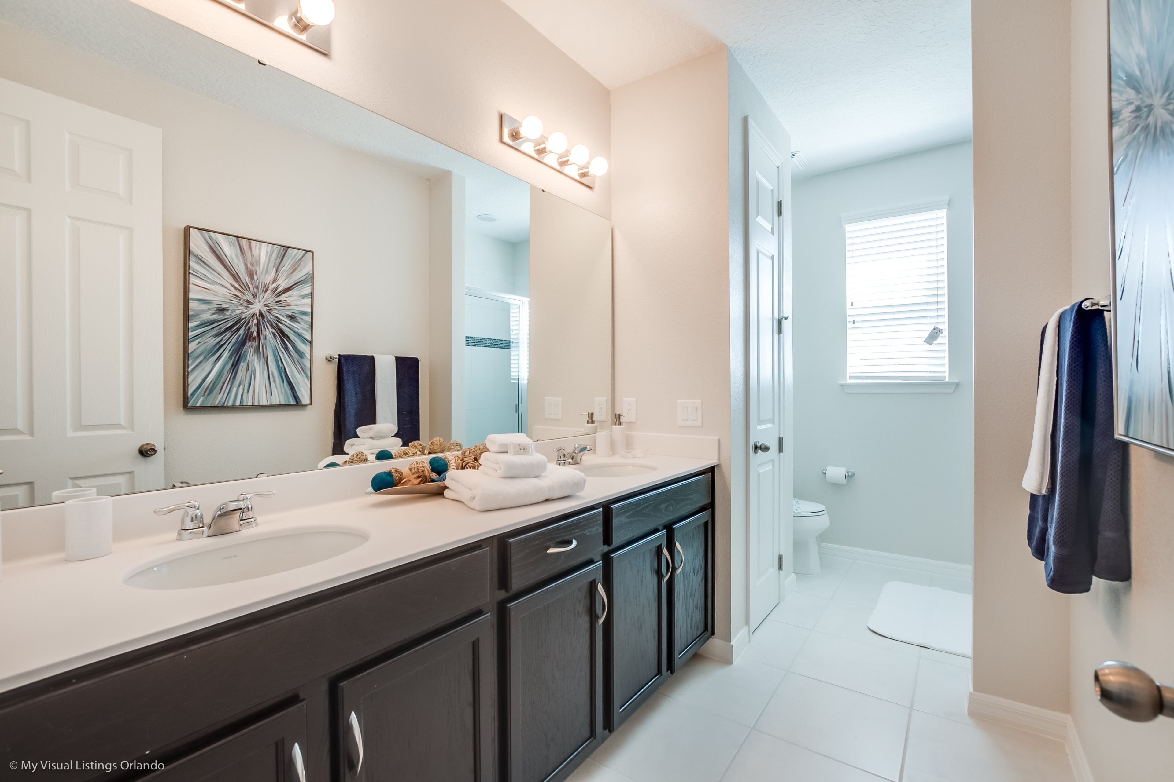A modern bathroom with two sinks, dark cabinets, a large mirror with lights, and a towel rack.