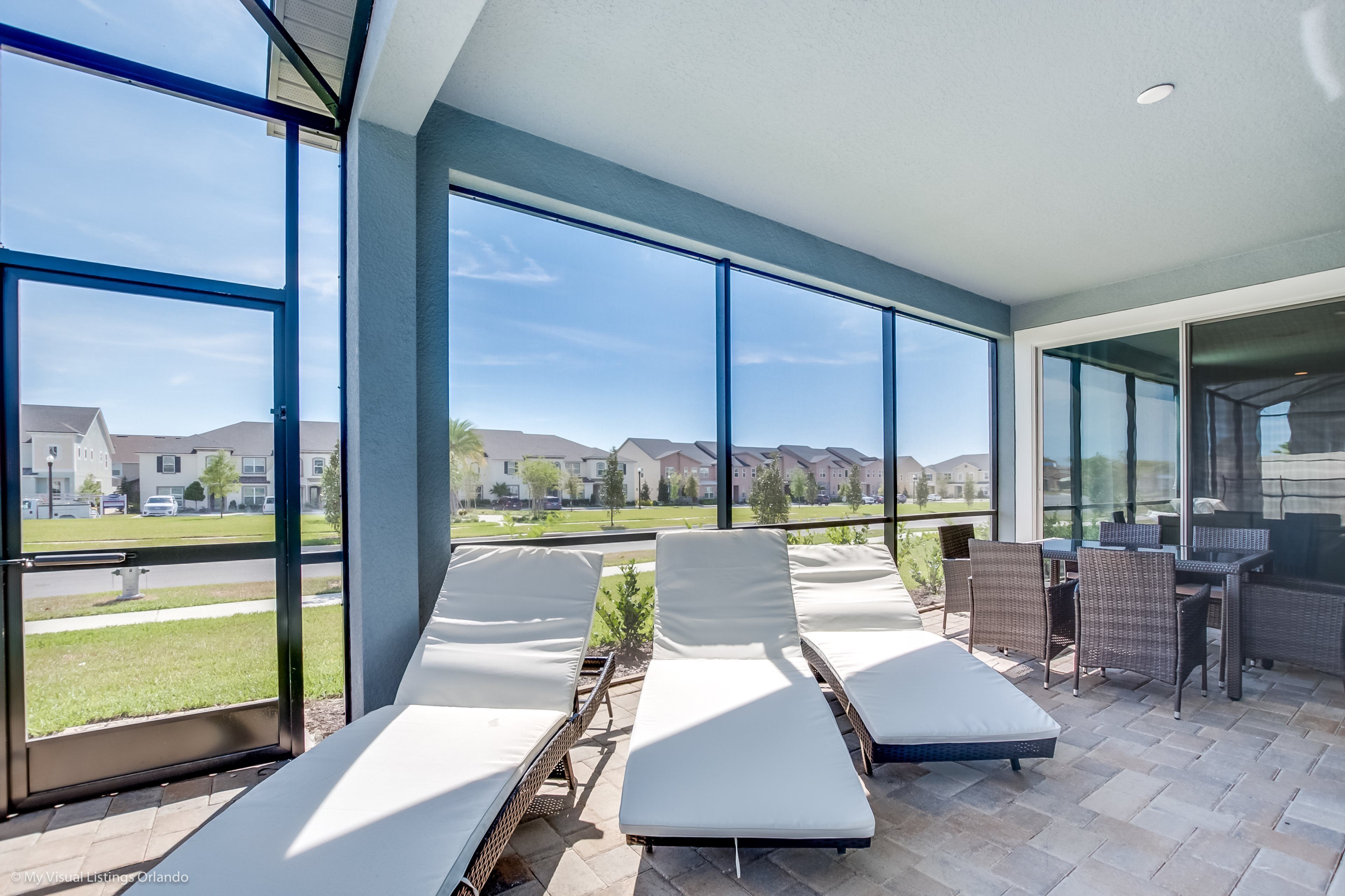 A sunlit outdoor lounge area with large windows, a patio table, and lounge chairs.