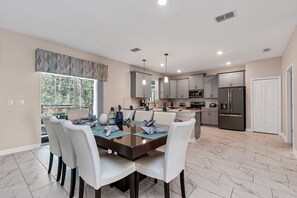 Una cocina moderna con armarios blancos, electrodomésticos de acero inoxidable y un comedor con mesa de vidrio y sillas blancas.
