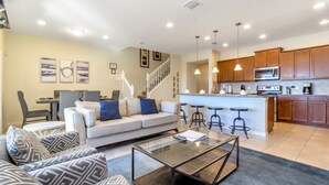 A modern living room with a sofa, coffee table, and bar stools. A kitchen with wooden cabinets and a microwave is visible in the background. - Lake View Villa (Kissimmee)