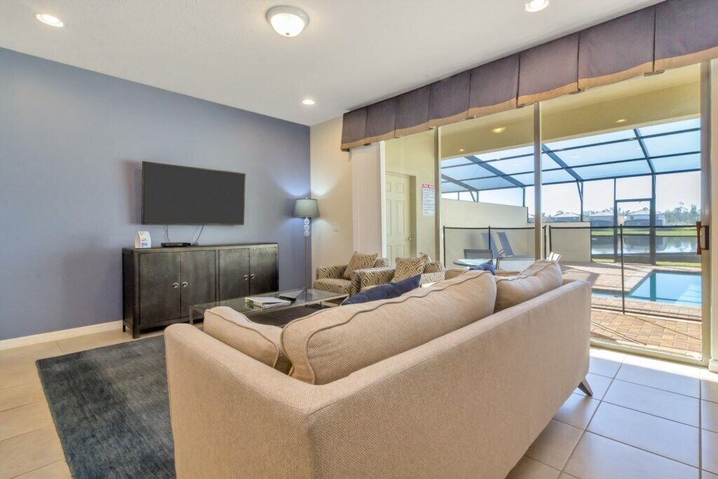 A modern living room with a large sofa, a flat-screen TV, a cabinet, and a glass door leading to a pool area.
