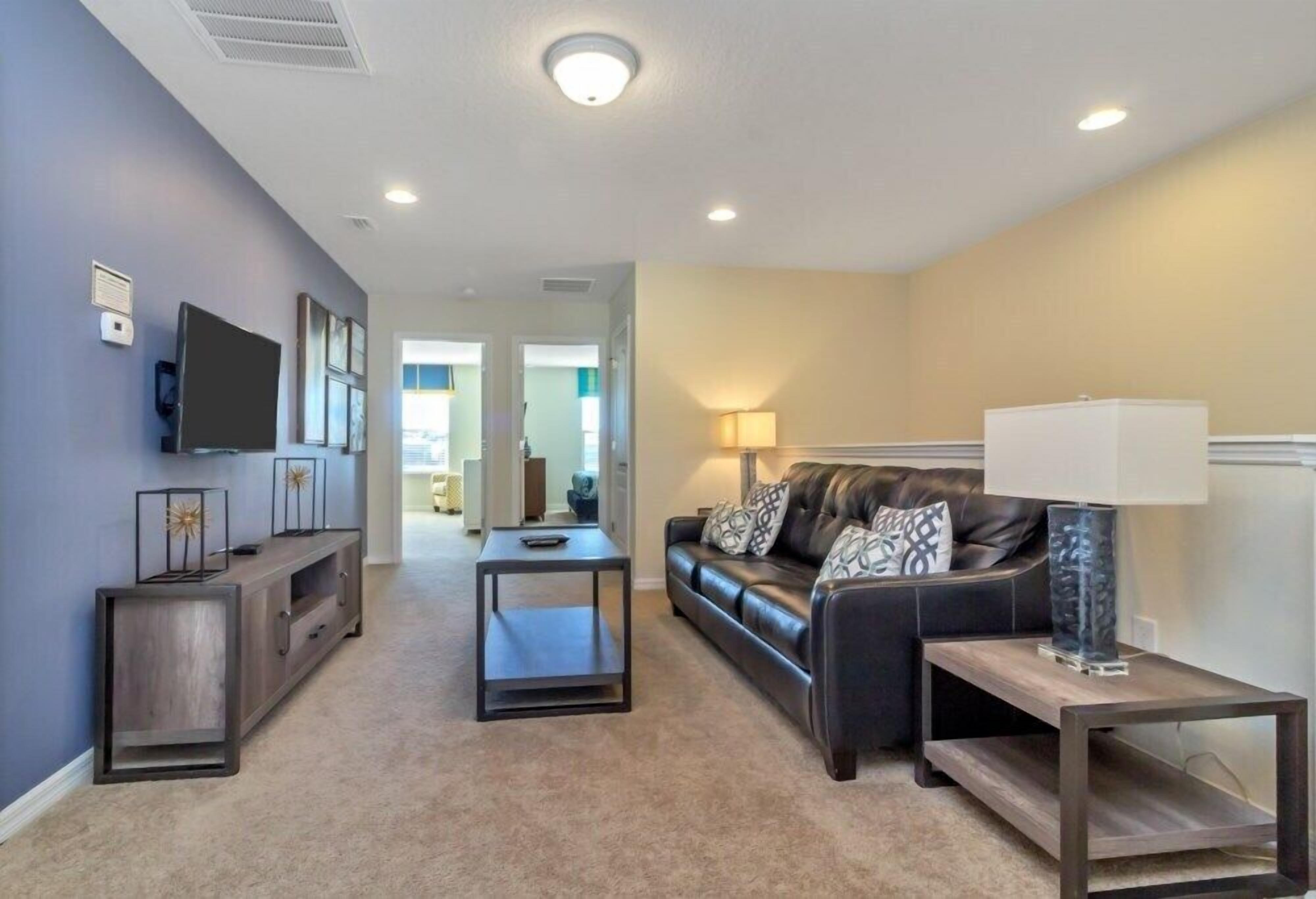 A living room with a dark leather sofa, a coffee table, a TV mounted on the wall, and a lamp on a side table.