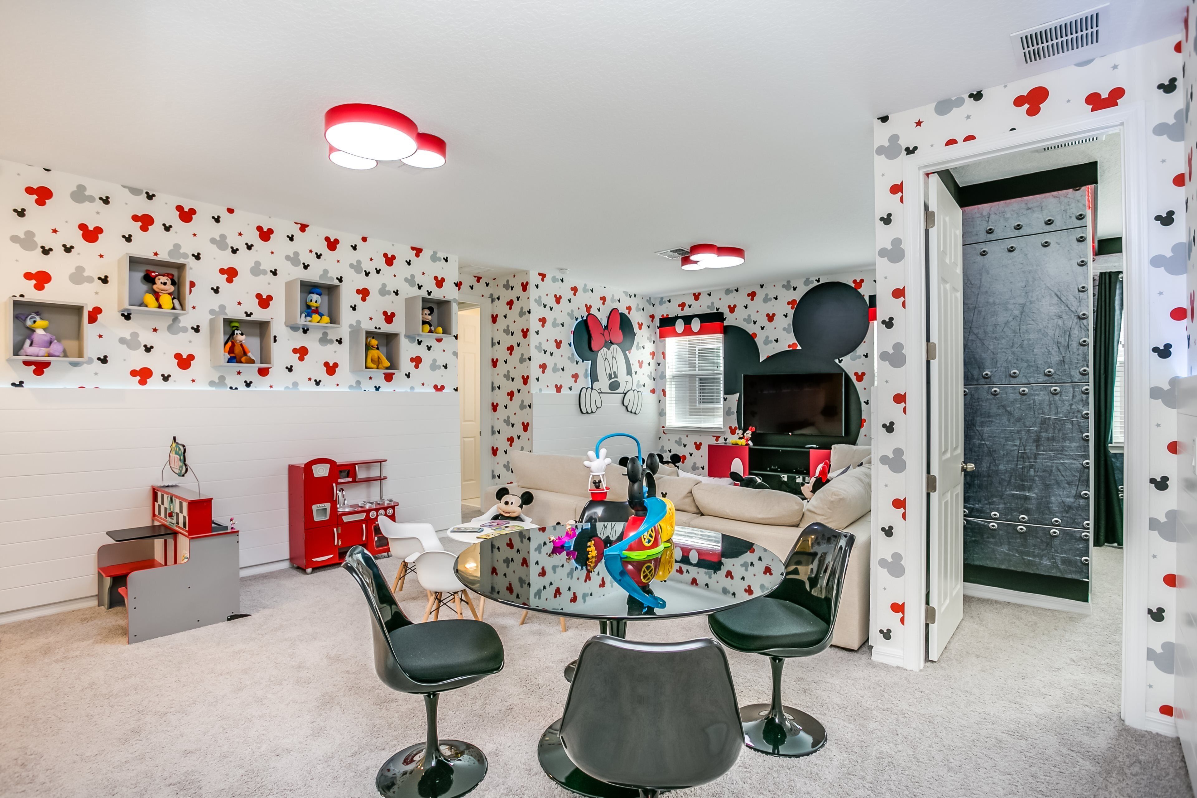 A room with a round glass table, black chairs, a red play area, and Mickey Mouse-themed decorations.