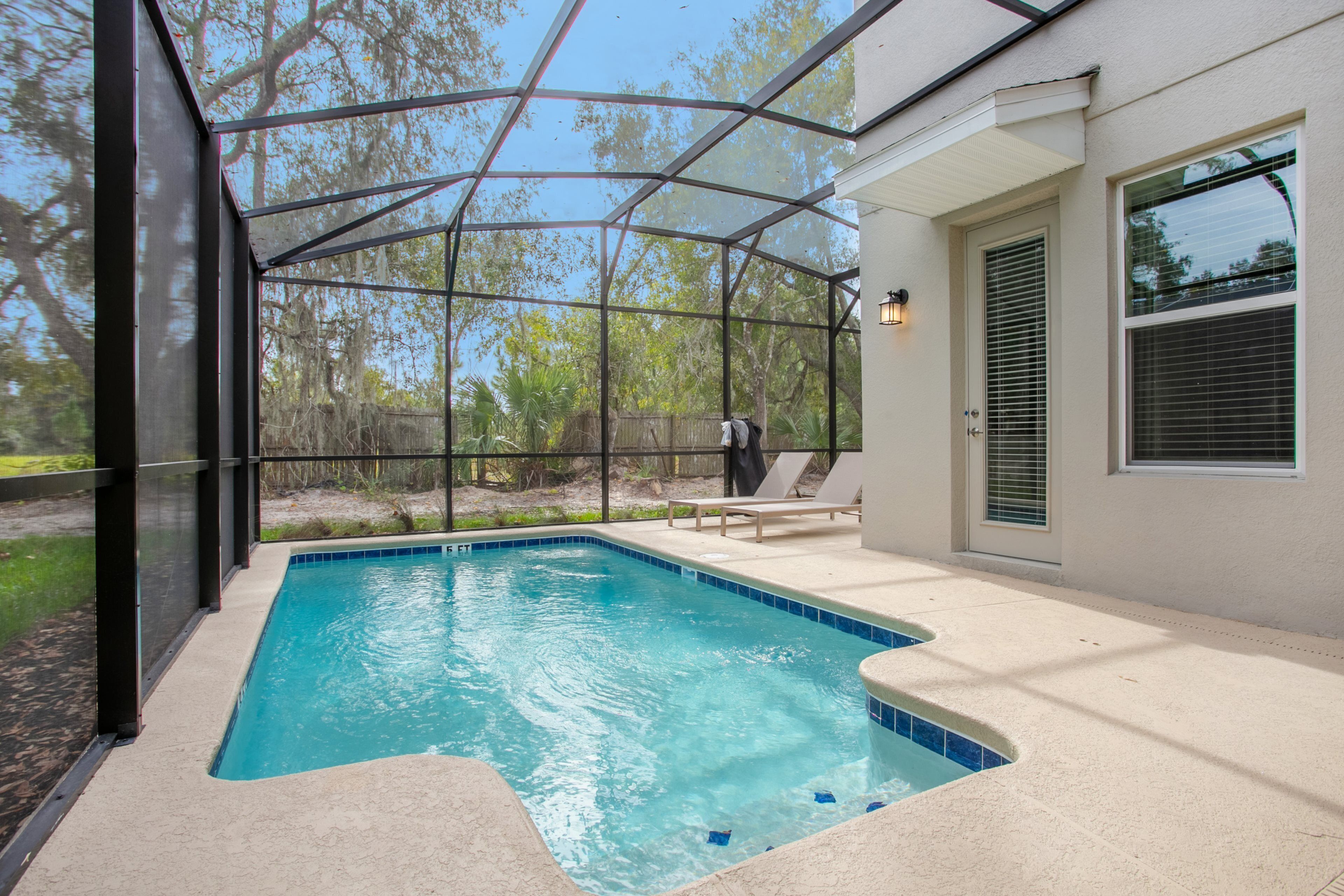 A swimming pool with a glass enclosure, a patio with lounge chairs, and a view of trees and a fence in the background.