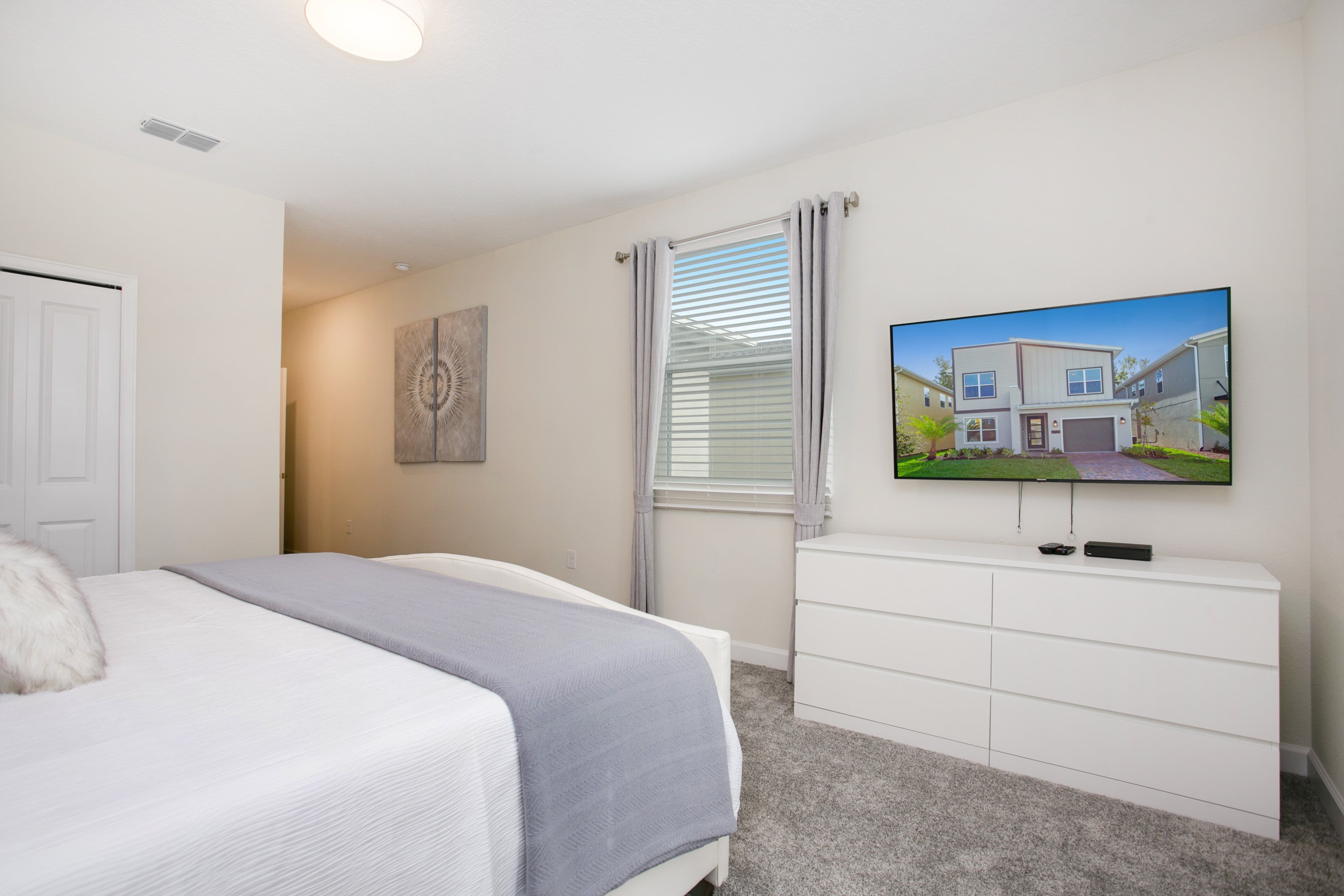 Une chambre à coucher comprenant un lit, une commode, un téléviseur fixé au mur et une fenêtre avec des stores.