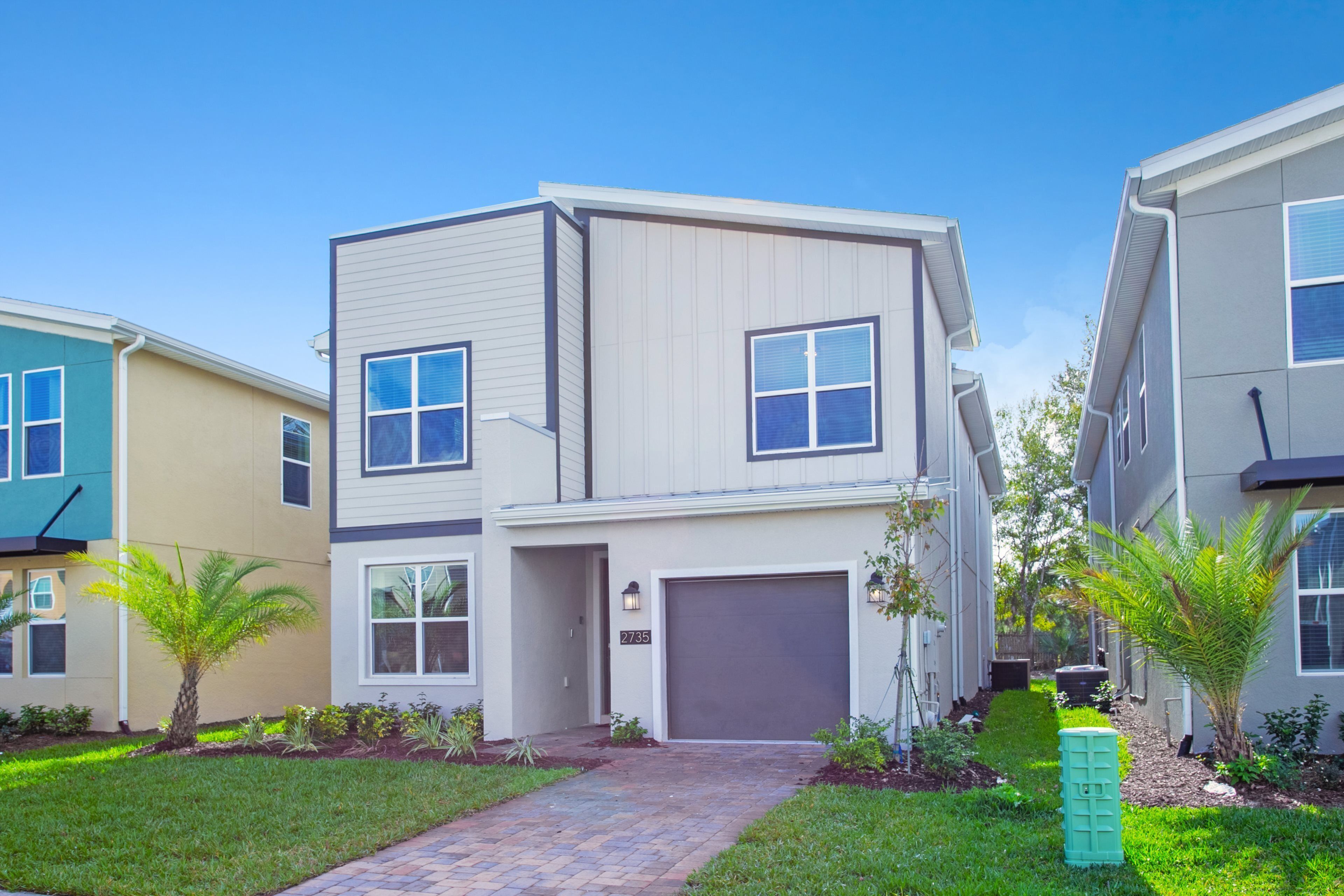 A modern two-story residential building with a paved driveway, a garage, and a small garden with palm trees.