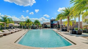 A swimming pool with lounge chairs and umbrellas, surrounded by palm trees and a modern building. - 3139brasillia (Kissimmee)