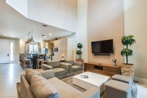 A spacious living area with a sofa, coffee table, and TV. There is a kitchen in the background with a dining area. - 3139brasillia (Kissimmee)