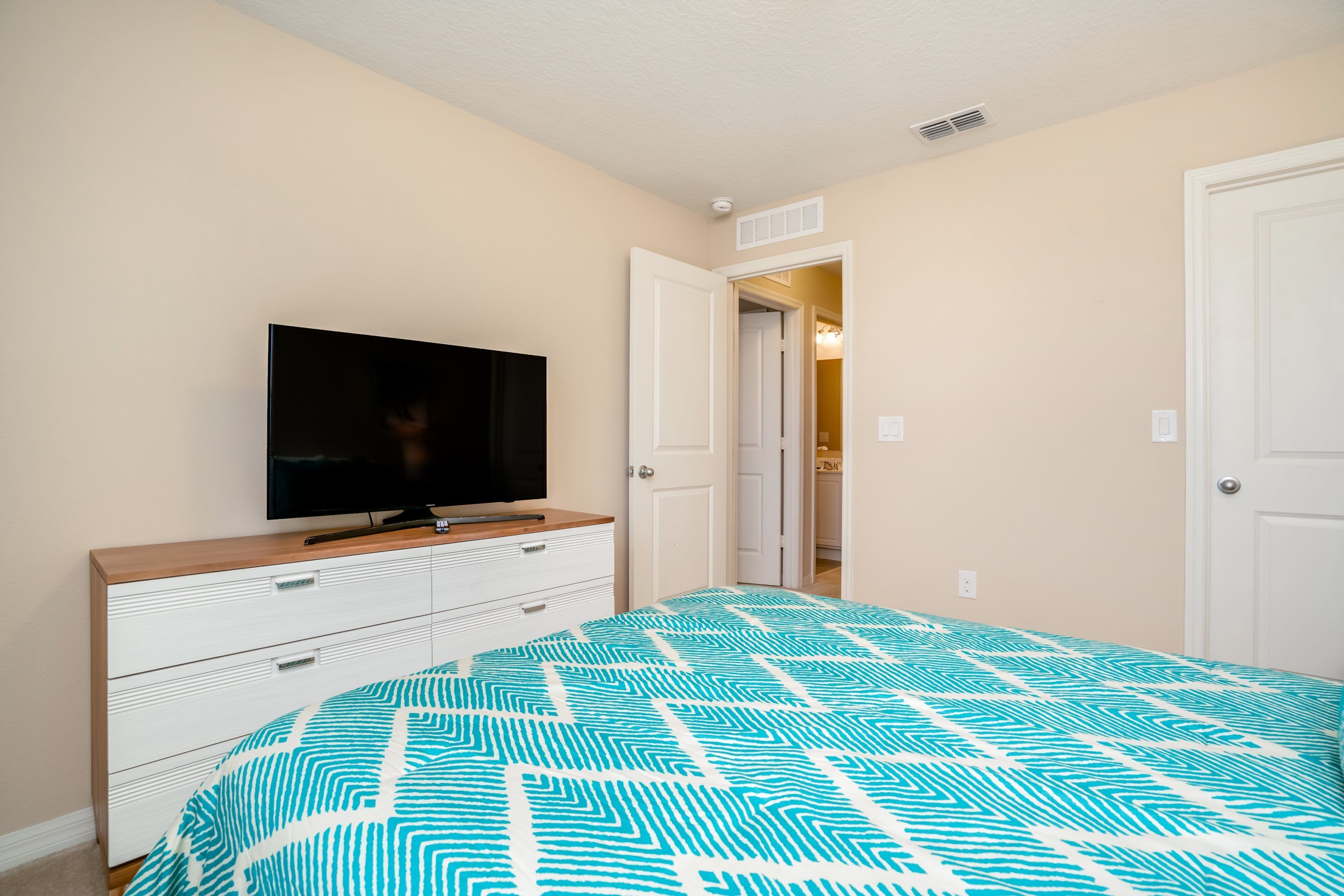 Un dormitorio con cama, televisor sobre una cómoda y puerta que da a otra habitación.