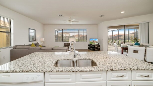 A modern kitchen with a granite countertop, stainless steel sink, and white cabinetry. There is a ceiling fan, a dining area with a table and chairs, and a large window with a view of the outdoors. - 2586rose (Davenport)