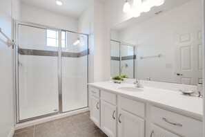 Une salle de bain moderne avec une douche à l’italienne en verre, un meuble-lavabo blanc doté d’un grand miroir et un sol carrelé de couleur beige.