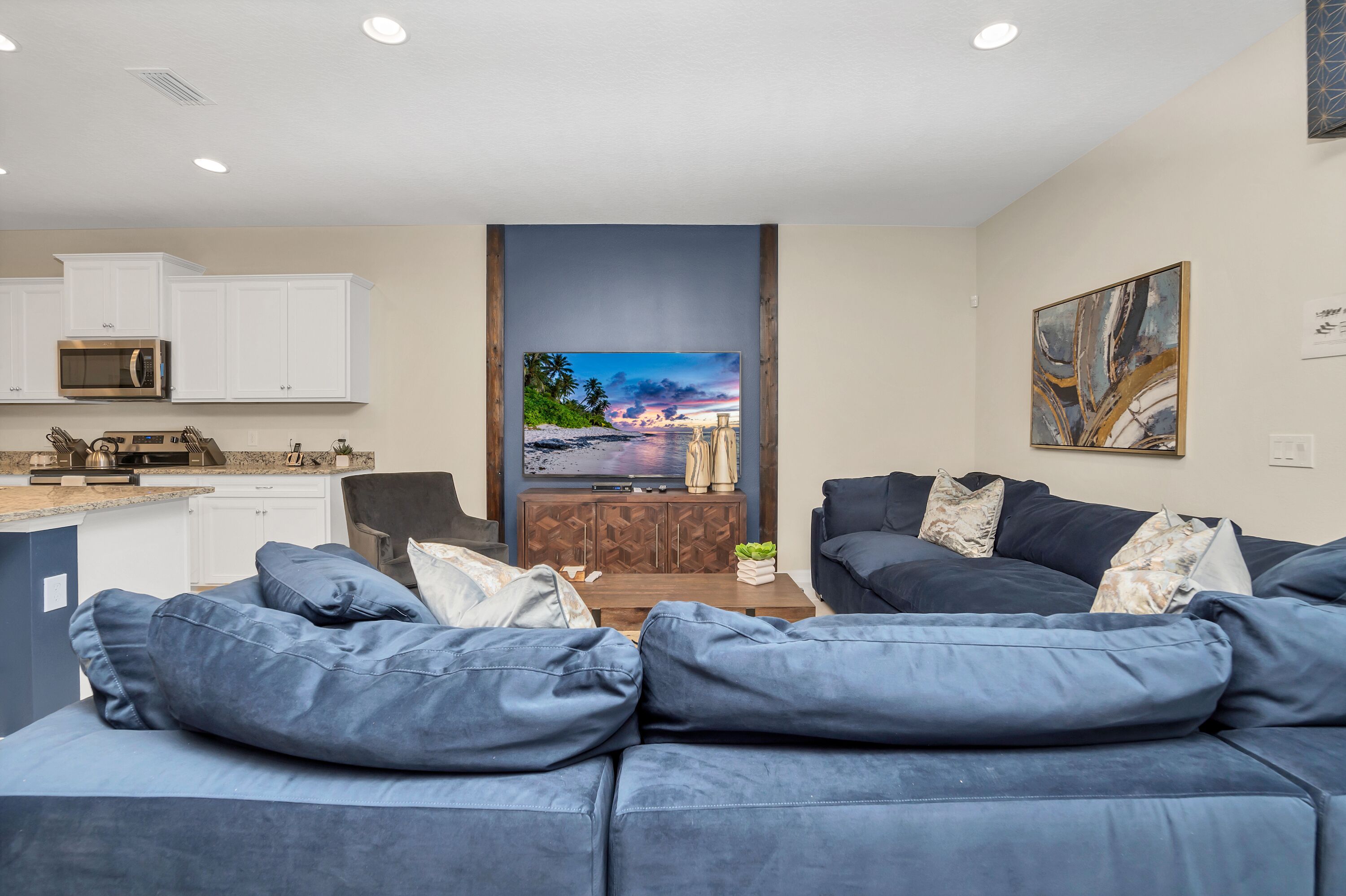 Una sala de estar moderna con un sofá grande azul, un televisor de pantalla plana y una zona de cocina con armarios blancos y un microondas.