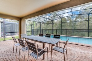 Une terrasse couverte avec une piscine, comprenant une table et des chaises, et de grandes fenêtres offrant une vue sur les arbres environnants.