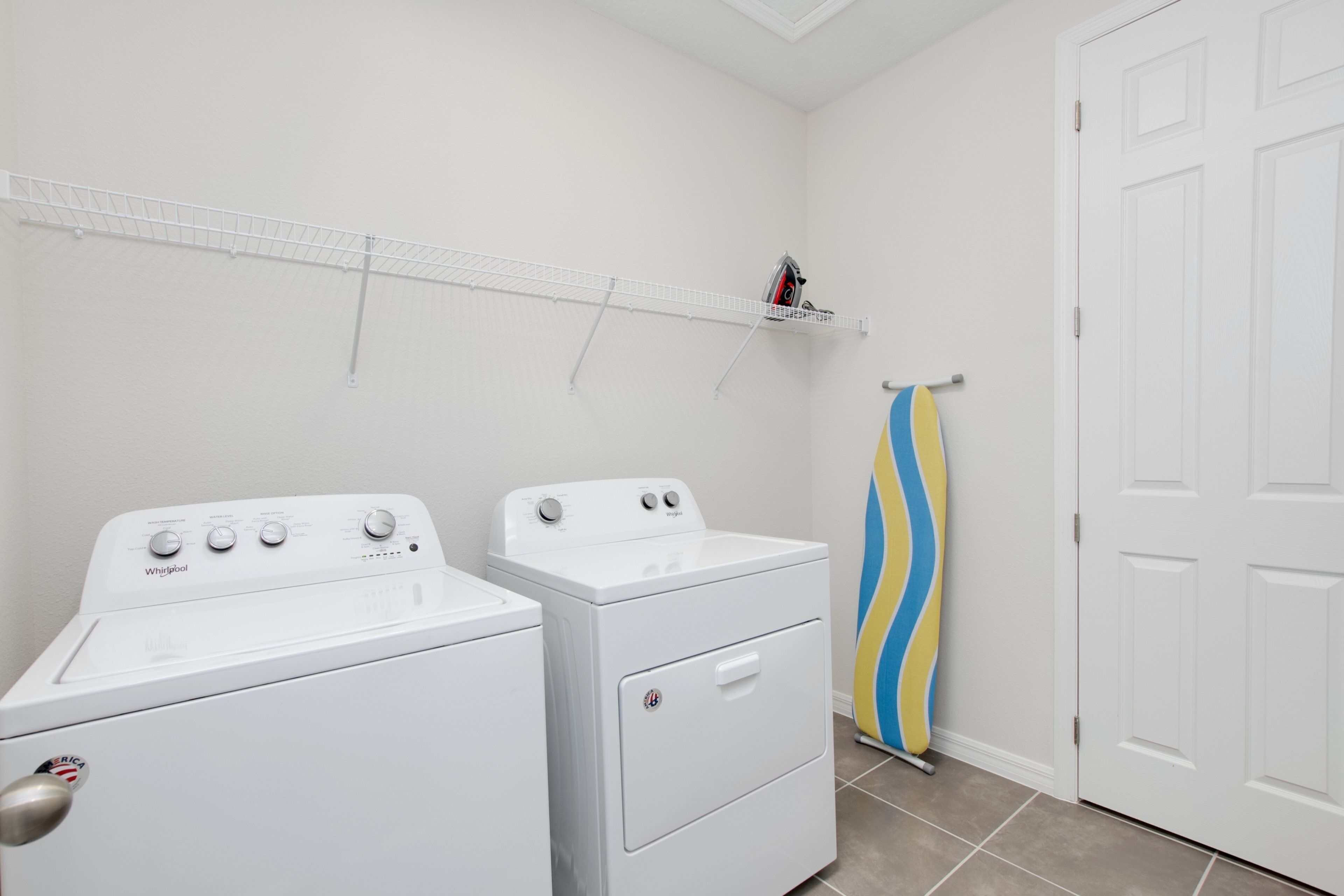 A laundry room with two white Whirlpool washers, a white shelf with a few items, and a door with a towel rack.