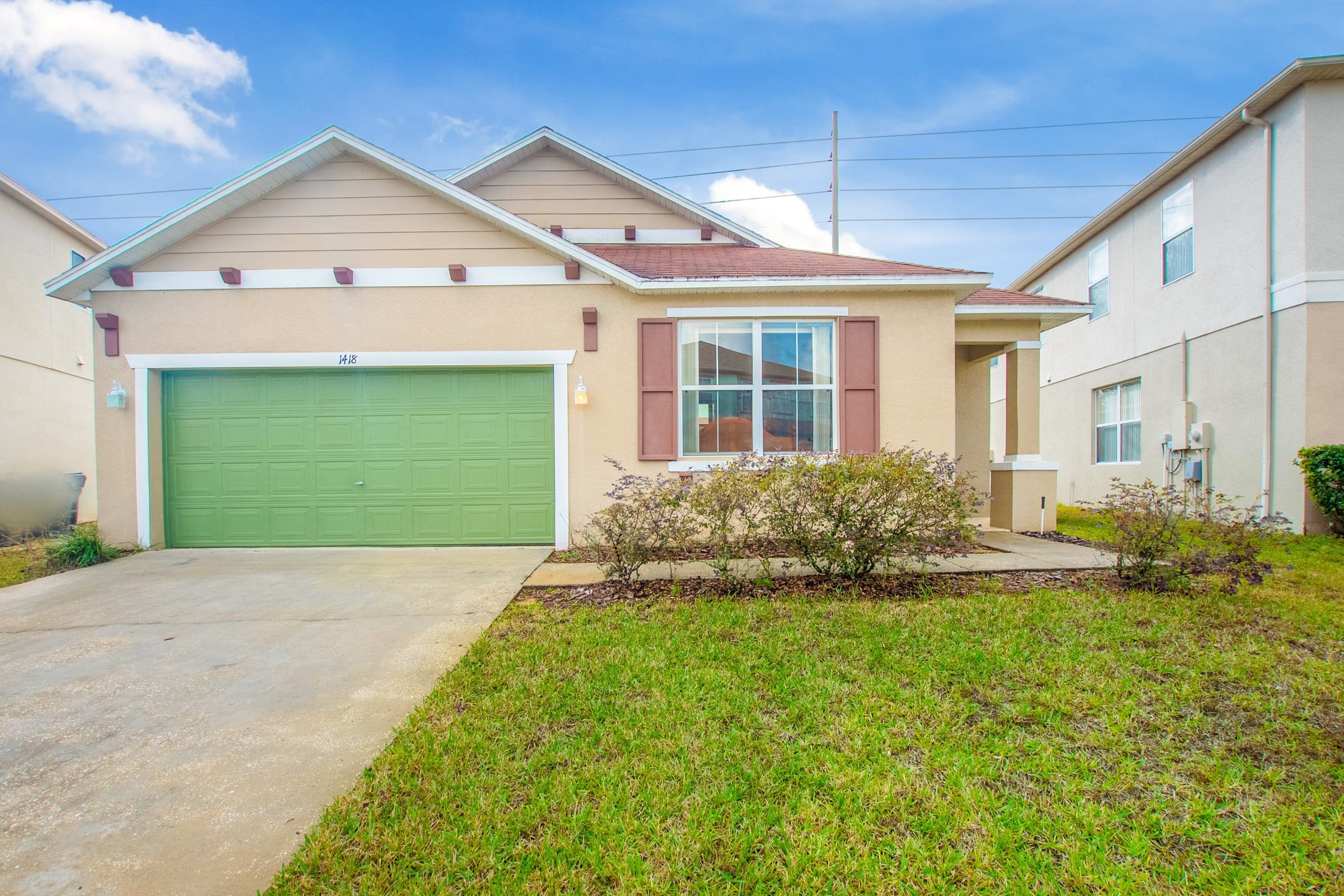 A two-story house with a green garage door, a small front yard with grass and a bush, and a clear blue sky.