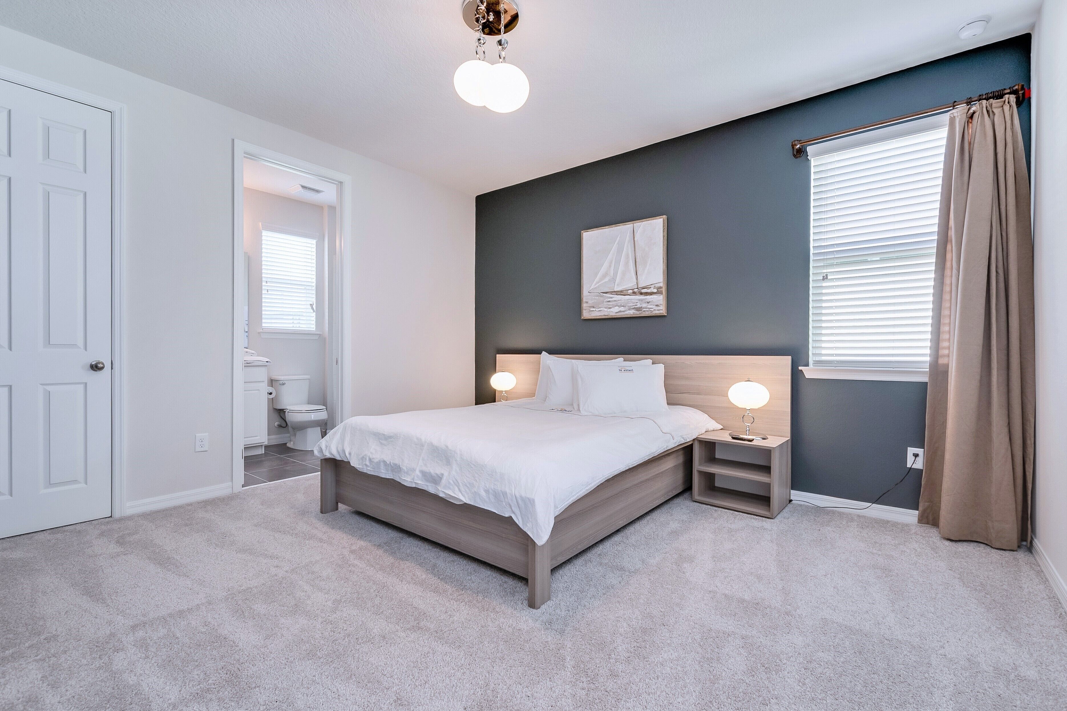 A bedroom with a bed, bedside tables, a nightstand, a wall-mounted lamp, a window with curtains, and a door leading to a bathroom.