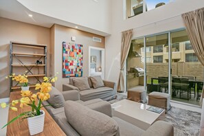 A modern living room with a sofa, coffee table, and potted plants. There is a colorful abstract painting on the wall and a sliding glass door leading to an outdoor area. - Modern Magic Village Resort Townhome (Kissimmee)