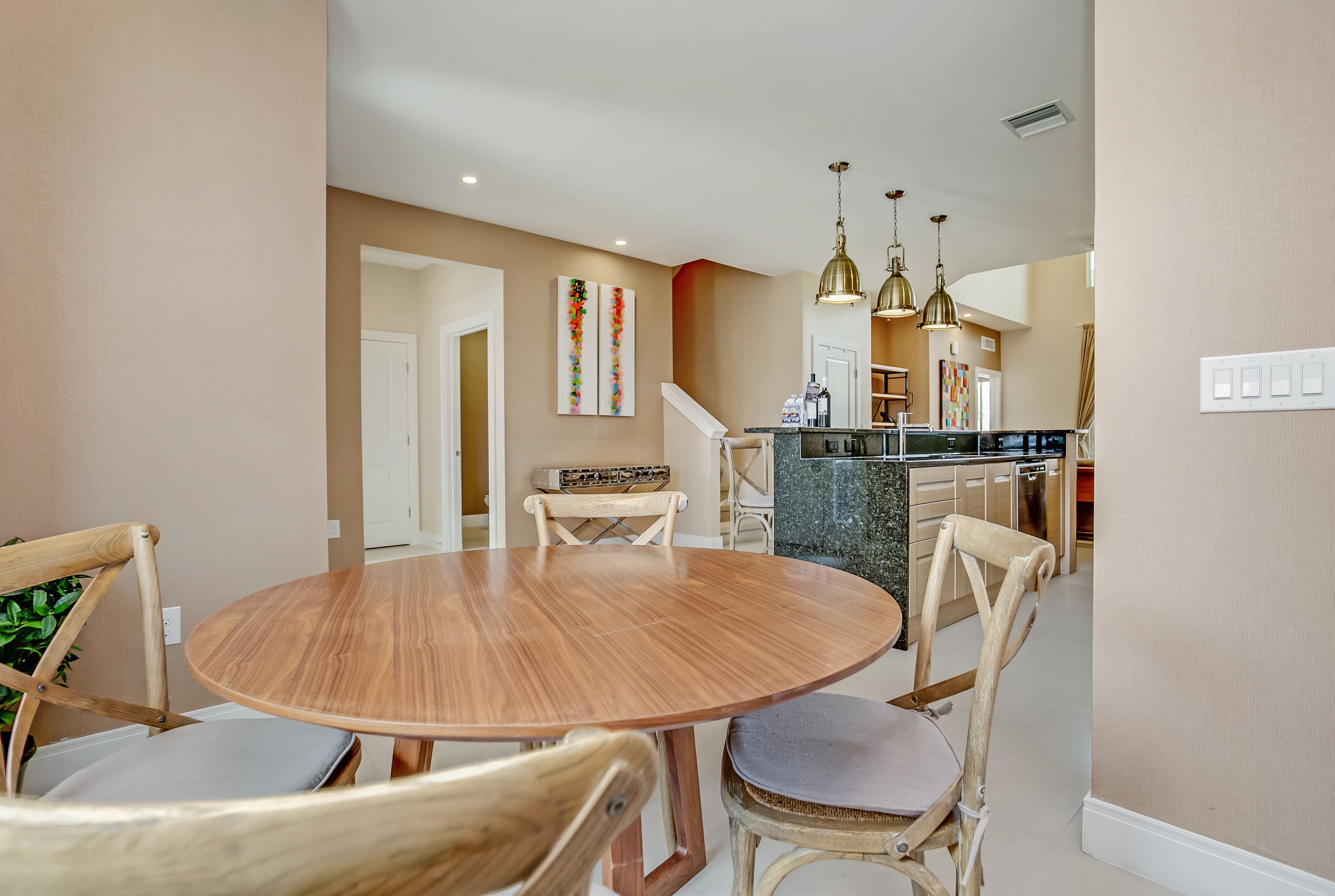 A dining area with a round wooden table and four chairs, a kitchen in the background, and pendant lighting.