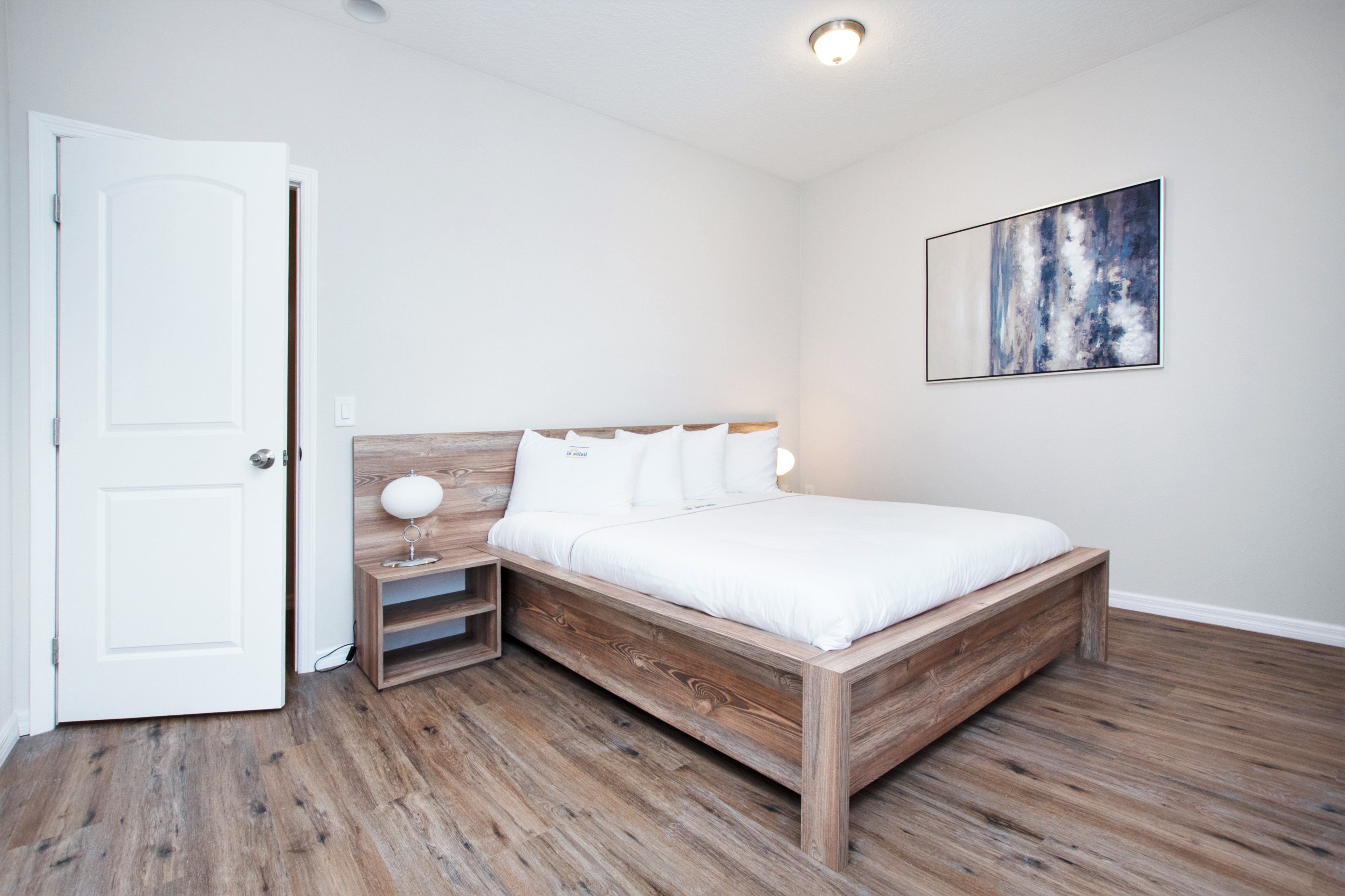 A bedroom with a wooden bed, white bedding, a bedside table with a lamp, and a painting on the wall.