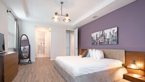 A bedroom with a large bed, a bedside table with a lamp, a dresser, a mirror, and a cityscape painting on the wall.