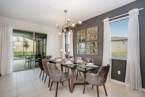 Comedor con mesa de vidrio, sillas grises, candelabro y vista a través de ventanas amplias.