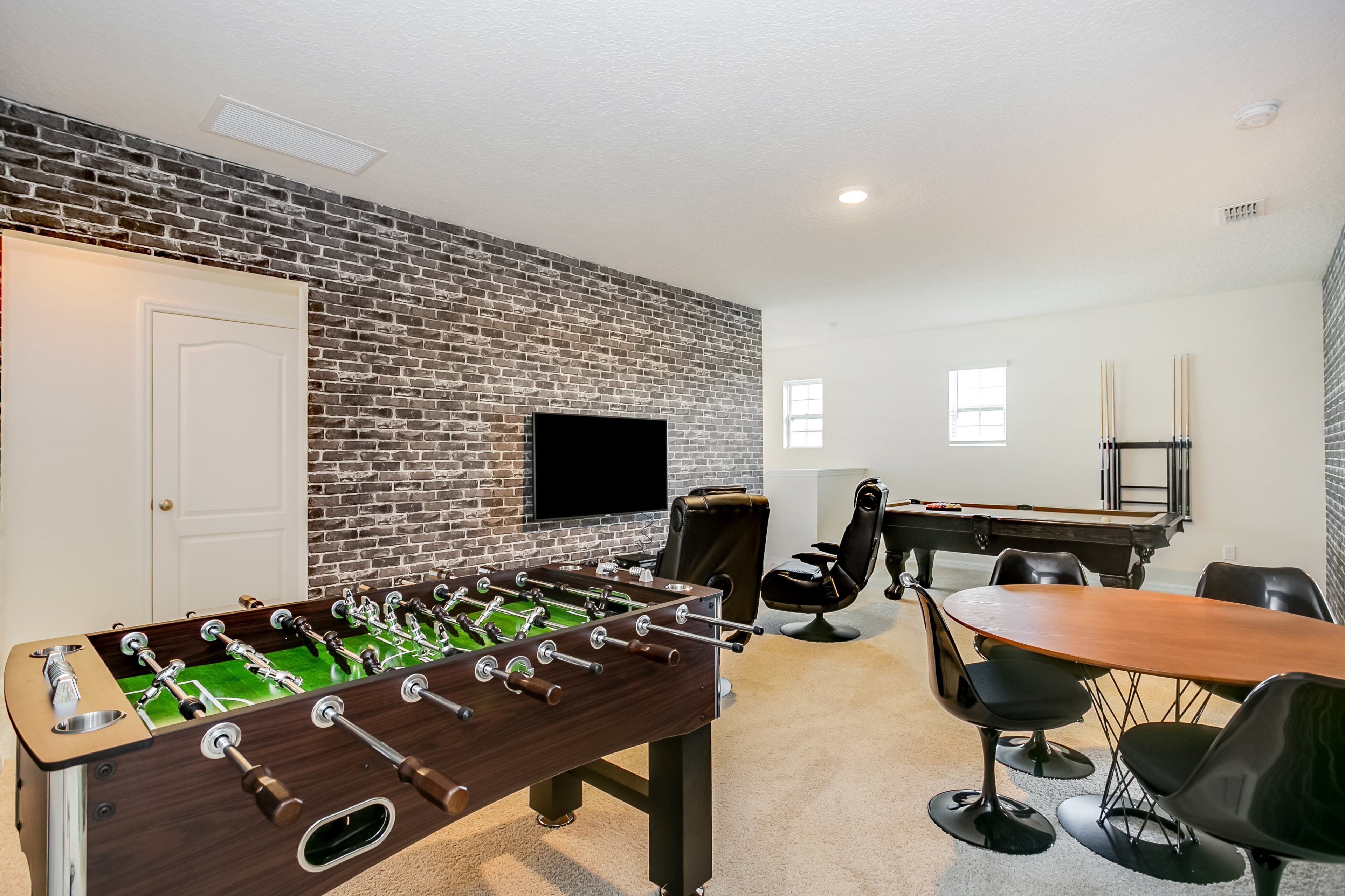 A room with a brick wall, a foosball table, a pool table, and a TV mounted on the wall.