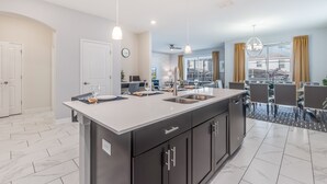 A modern kitchen with a central island, a dining area with a high table and chairs, and a view of the outside through large windows. - 1559muligan (Davenport)