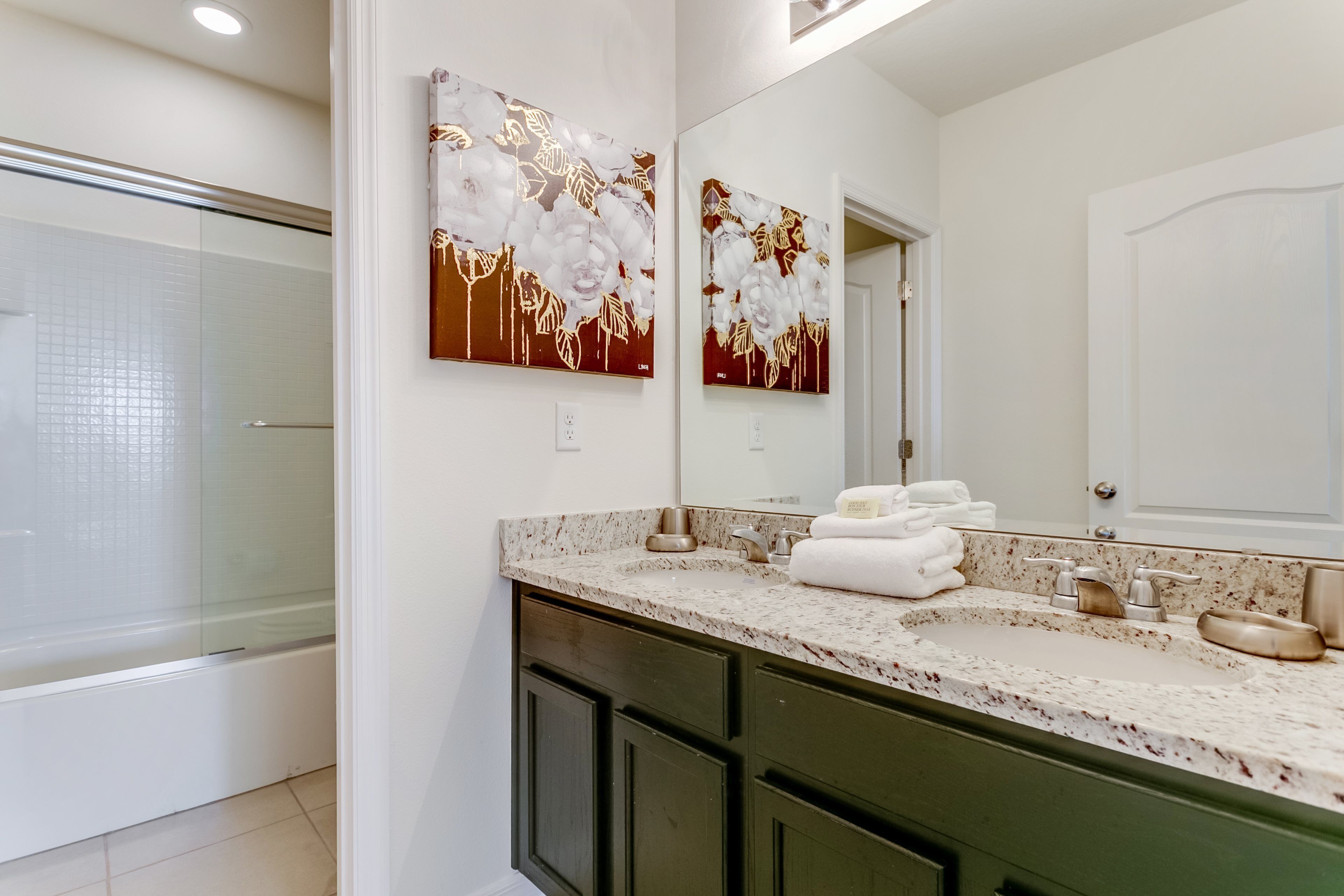 A modern bathroom with a large mirror, two wall-mounted artwork pieces, a granite countertop, and a glass shower enclosure.