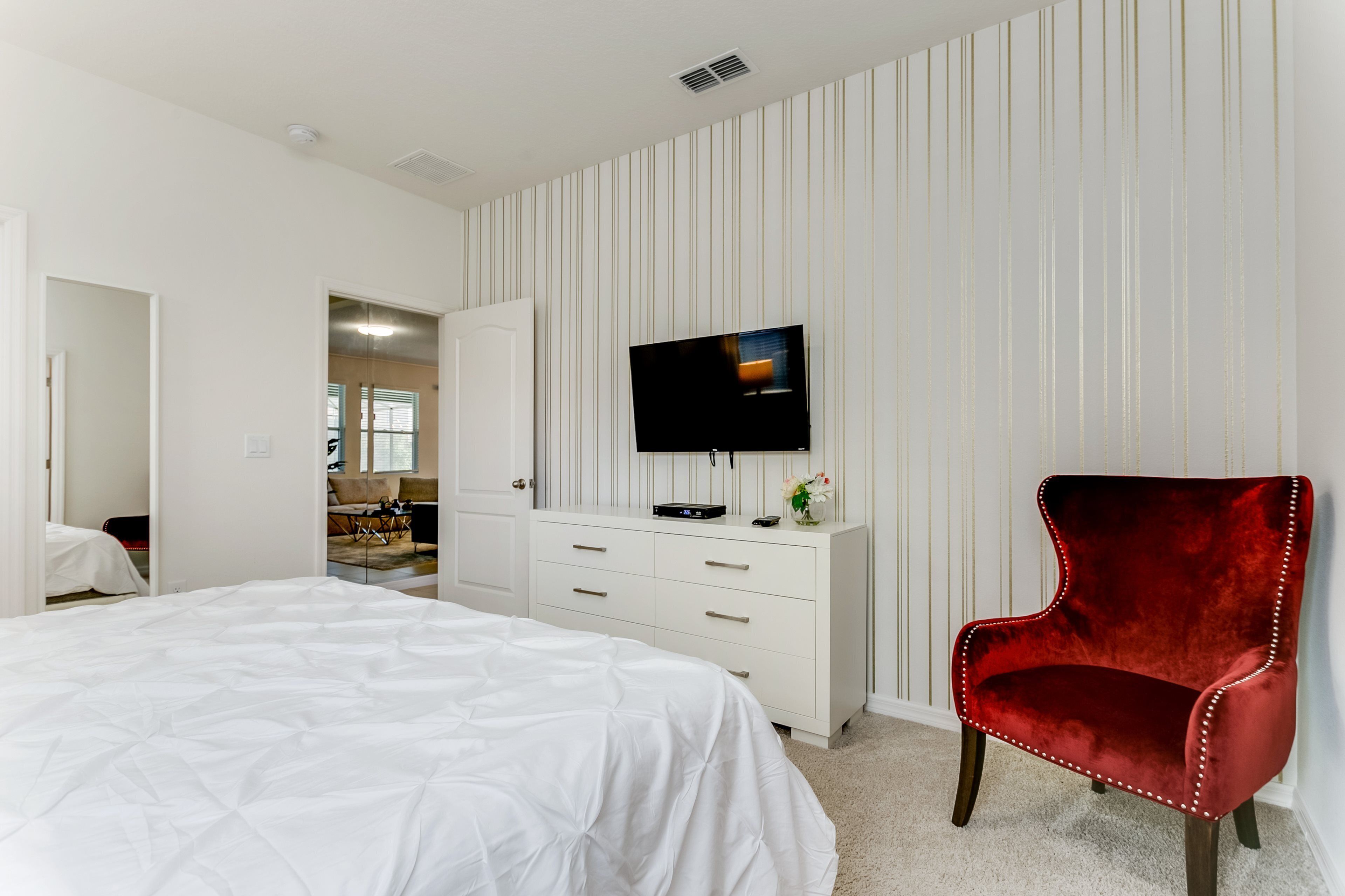 A bedroom with a large bed, a red chair, a TV on a white dresser, and a striped wall.
