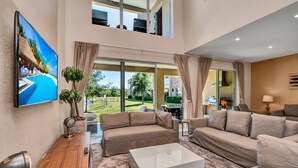 A spacious living room with a large flat-screen TV, beige sofas, a coffee table, and a view of a garden through large windows. - Modern Magic Village Resort Townhouse 7620 (Kissimmee)