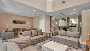 A spacious living area with a neutral color scheme, featuring a large sectional sofa, a coffee table, and a dining area with a kitchen in the background. - Modern Magic Village Resort Townhouse 7620 (Kissimmee)
