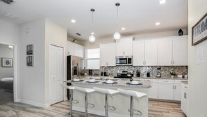 A modern kitchen with white cabinets, a central island with bar stools, stainless steel appliances, and pendant lighting. - 5 Bed 4 Ba Villa with Game Room, Pool & Spa (Kissimmee)