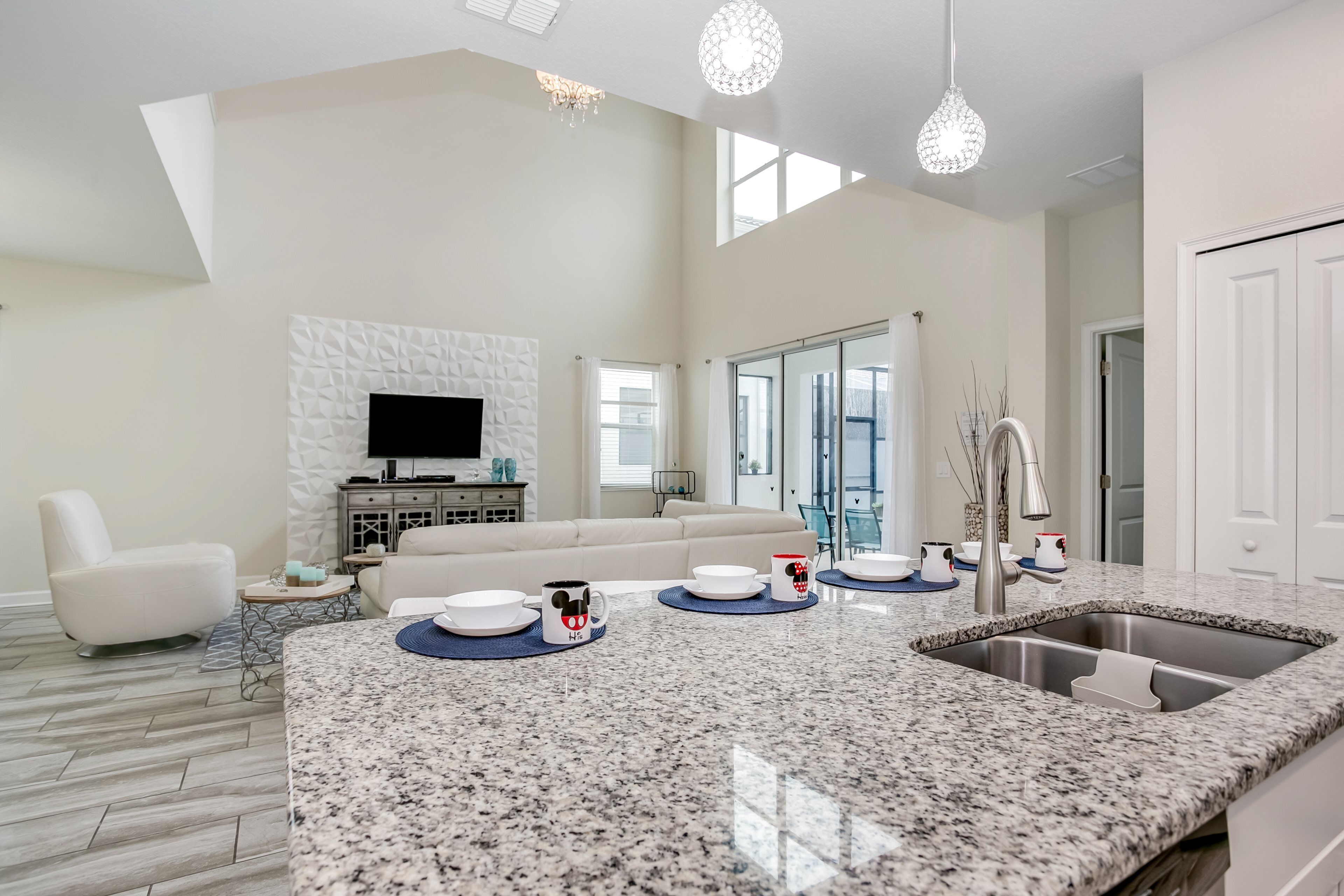 A modern kitchen with granite countertops, a stainless steel sink, and a dining area with a white sofa and a TV.