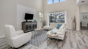 A spacious living room with a white leather sofa, a flat-screen TV mounted on a textured wall, and a large window offering a view of a greenhouse. - 5 Bed 4 Ba Villa with Game Room, Pool & Spa (Kissimmee)