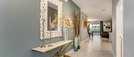 A hallway with a marble floor, a white ornate mirror, a gold console table, and tall vases with dried branches.