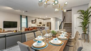 A modern dining area with a long table set for six, a kitchen in the background, and a staircase leading to another level.