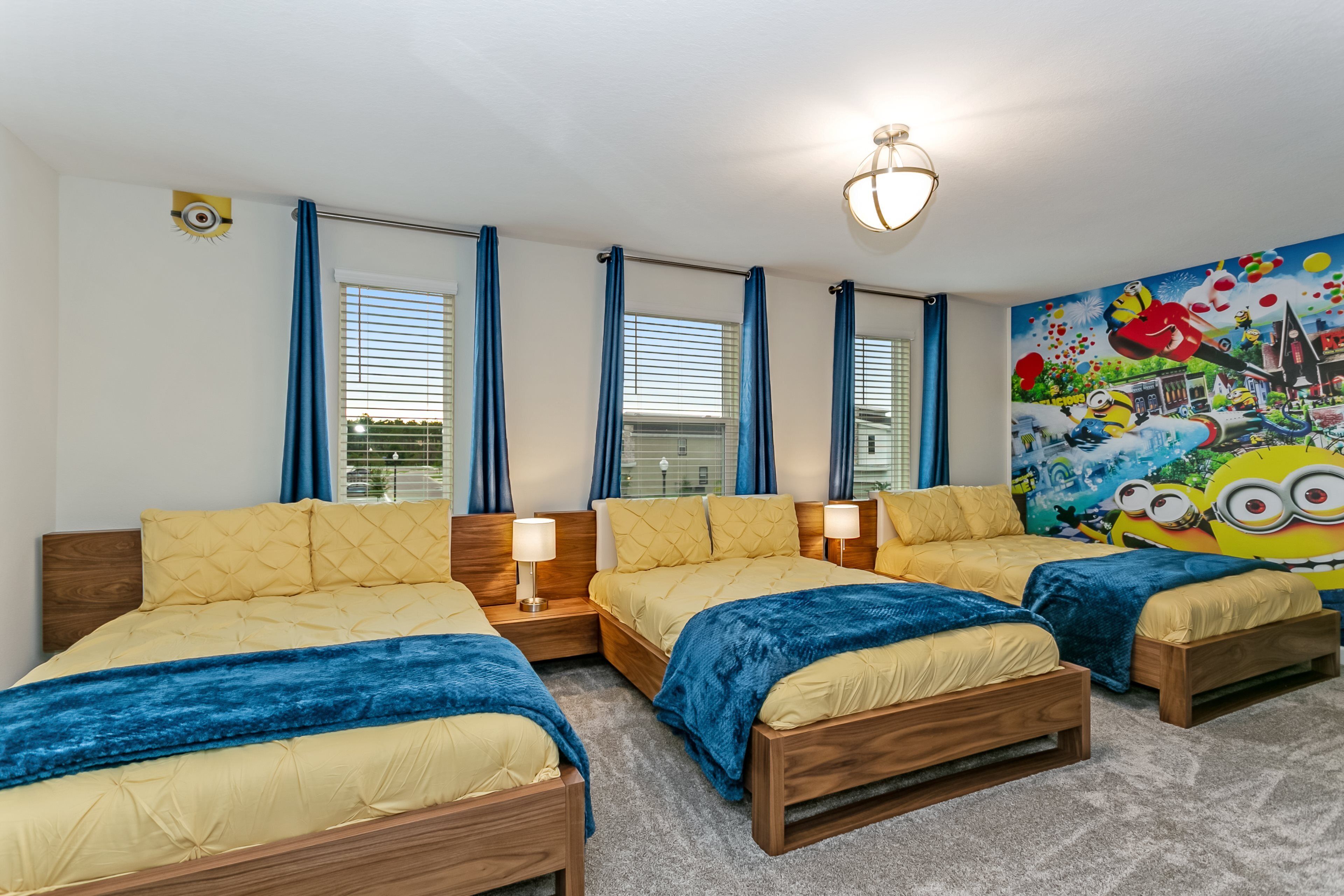 A hotel room with three beds, a large window with blinds, a Minion-themed mural, and a ceiling light fixture.